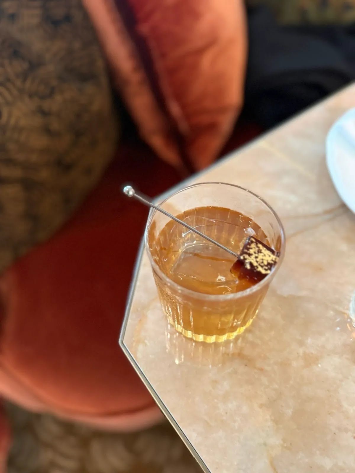 An amber cocktail over a large ice cube on a marble table beside red seating